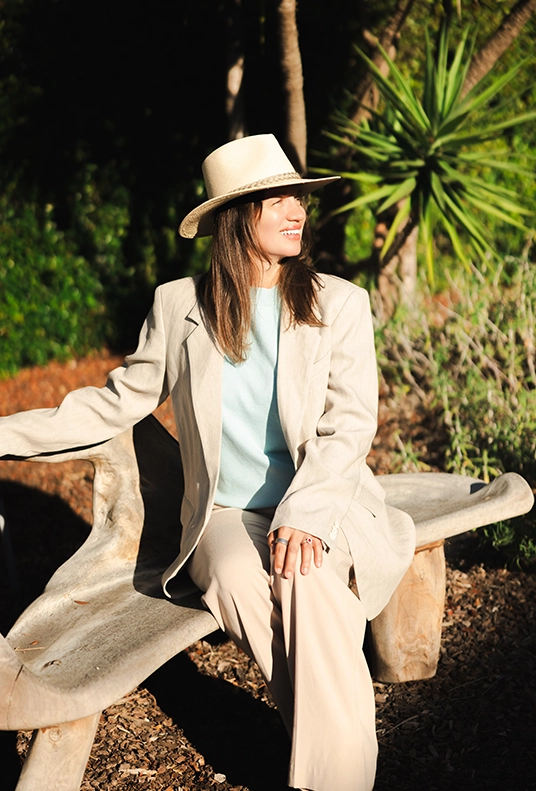 DHC Insights founder seated outdoors wearing a light blazer and wide-brim hat.